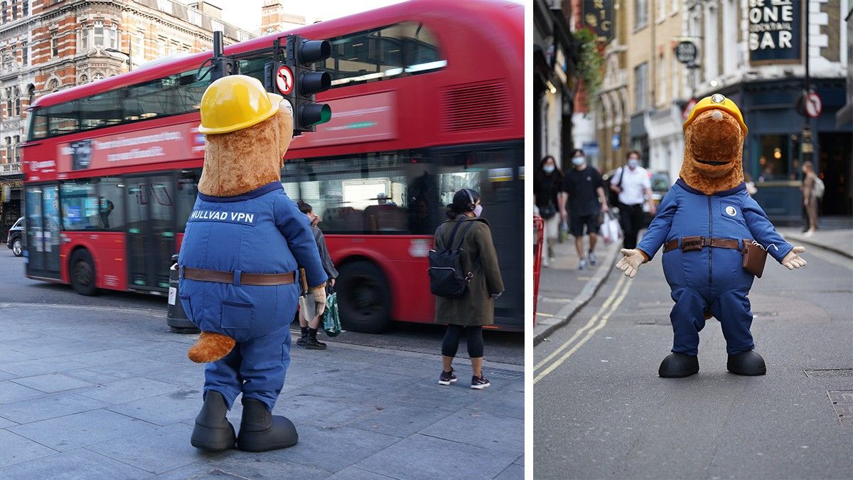 Mole mascot in a yellow hardhat and blue jumpsuit with MULLVAD VPN text, standing next to a red double-decker bus on a city street.